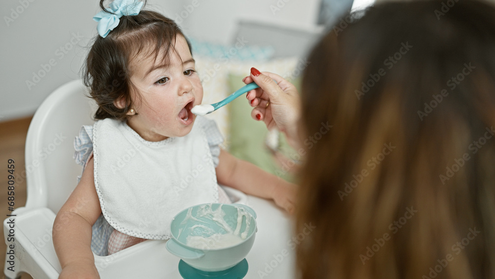 Mother and daughter together, loving family lifestyle, casually sitting at home during baby's lunch time, holding spoon and bib, concentrating on eating in the living room interior. Mother and daughter together, loving family lifestyle, casually sitting at home during baby's lunch time, holding spoon and bib, concentrating on eating in the living room interior.