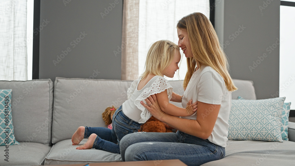 Relaxed caucasian mother and her little girl confidently sharing smiles indoors, comfortably sitting on living room sofa encapsulating family lifestyle.