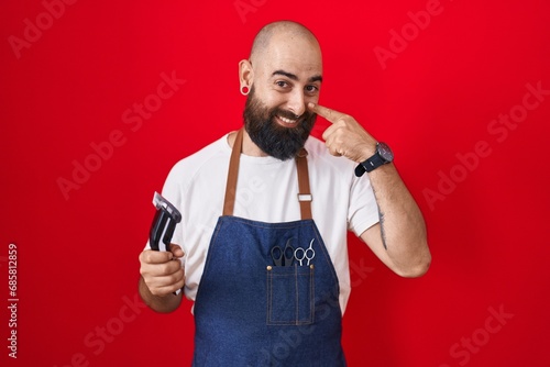 Young hispanic man with beard and tattoos wearing barber apron holding razor pointing with hand finger to face and nose, smiling cheerful. beauty concept