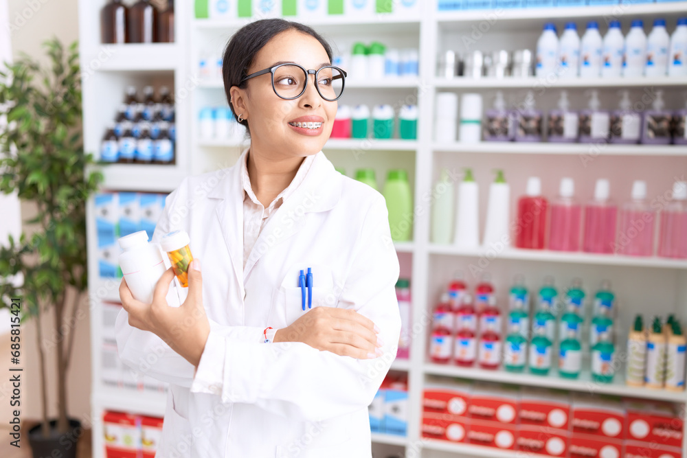 Young arab woman pharmacist holding pills bottles standing with arms crossed gesture at pharmacy