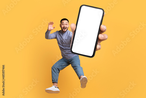 Asian man holding large cellphone blank screen leaping in studio