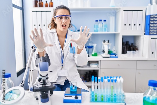 Young hispanic woman working at scientist laboratory afraid and terrified with fear expression stop gesture with hands  shouting in shock. panic concept.