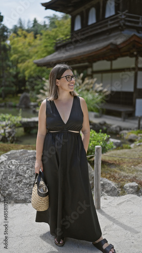 Cheerful young hispanic woman, effortlessly beautiful with glasses, confidently posing and joyfully smiling while exploring the serene surroundings of ginkaku-ji temple, kyoto, japan