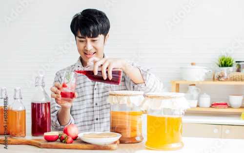 Portrait handsome man smiling with fresh and happiness, holding glass of Kombucha healthy fermented probiotic tea drinks with ingredients red apples, strawberries, mushroom scoby in kitchen at home.