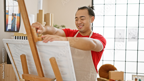 Confident young chinese man with stylish pigtail hairstyle, joyfully putting canvas on easel in indoor art studio, prepping for an exciting painting lesson, radiating a heartwarming smile.