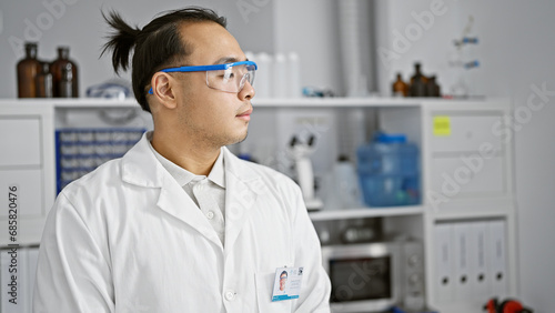 Serious young chinese man  a lab scientist in safety glasses  deep in concentration on medical research at a bustling  professional chemistry lab.