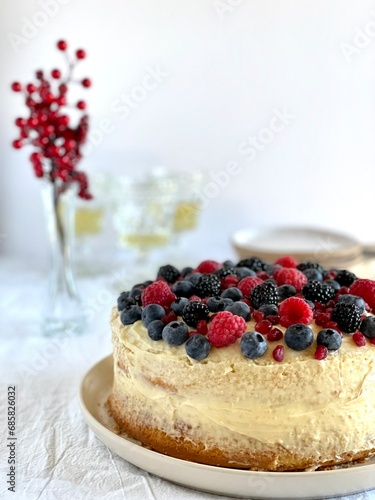 Homemade vanilla cake with buttercream frosting and fresh berries on a white surface with glasses of white wine in the background. Christmas cake. Copy space