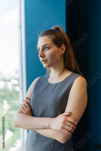 Free Download Dreamy lady with crossed arms looks away from camera posing for photo near window. Young brunette woman with high ponytail looks in window - GFXLoad