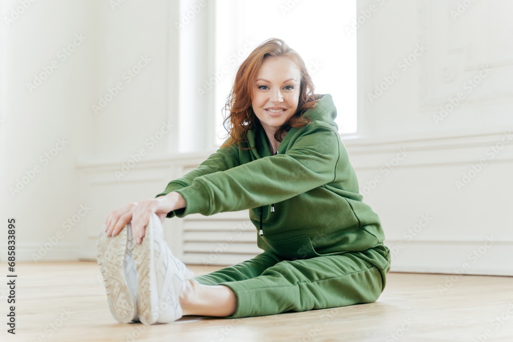 Fit woman in green tracksuit stretching, touching toes, relaxed fitness routine in a bright room with wooden floor Fit woman in green tracksuit stretching, touching toes, relaxed fitness routine in a bright room with wooden floor