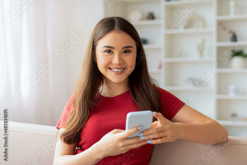 Smiling Asian Woman With Smartphone In Hands Sitting On Couch At Home
