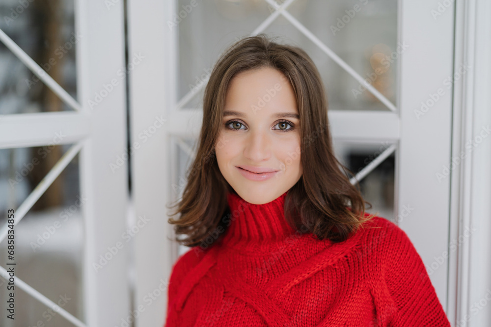 Smiling woman in a red turtleneck sweater, looking cozy and stylish, with a clear window and winter scene behind Smiling woman in a red turtleneck sweater, looking cozy and stylish, with a clear window and winter scene behind