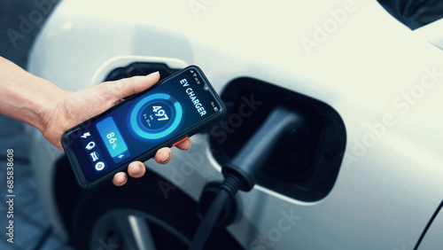 Businessman holding smartphone display battery status interface by smart EV mobile application while EV car recharging electricity from charging station in car park. Peruse