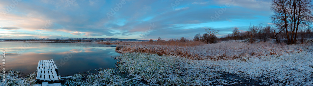 Panorama of the sunrise over the winter river. The sun rises over the lake. Autumn frost. The frozen river is covered with thin ice, the grass glistens with frost. Panorama of the sunrise over the winter river. The sun rises over the lake. Autumn frost. The frozen river is covered with thin ice, the grass glistens with frost.