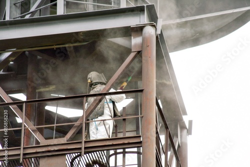 Sand blasting process. Industrial worker in protective uniform cleaning surface of big steel construction before painting on high altitude.
