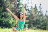 Young cheerful brunette girl in green sportswear sitting in lotus pose in green grass outdoors looks at camera smiles riding hands happily. Fit caucasian young woman trains at park, sunny day