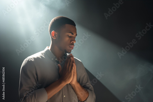 Handsome young African ethnic man praying to god with his eyes closed and clasped hands - profile side angle - God's rays of light shining down - Ethnic diversity and religion concept