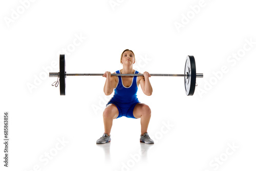 Concentrated strong young woman, athlete training doing squats, lifting heavy barbell against white background. Concept of sport, strength, gym, healthy lifestyle, power and endurance, weightlifting.