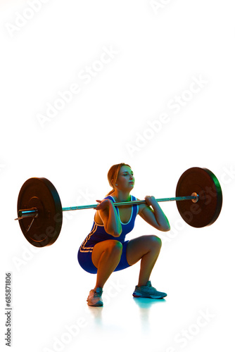 Young woman, athlete in motion, training, lifting heavy weights, barbell against white background in neon light. Concept of sport, strength, gym, healthy lifestyle, power and endurance, weightlifting.