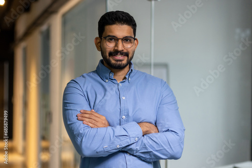 Businessman with crossed arms smiling and looking at camera, portrait of happy male worker inside office, boss in glasses and shirt, happy with achievement results, proud and confident.