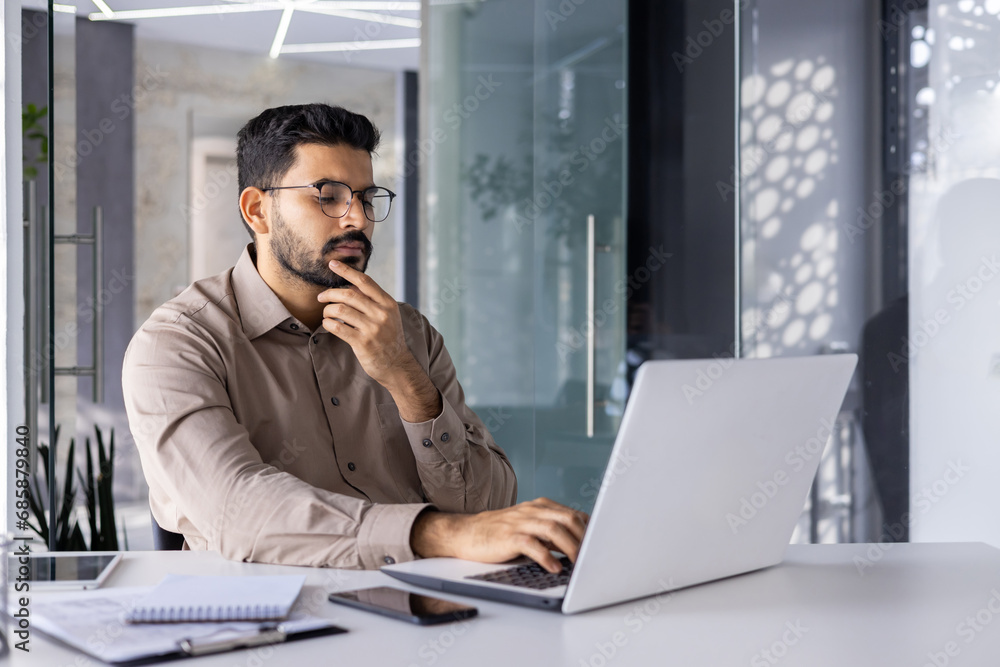 Man thinking inside office at workplace with laptop, businessman preparing financial report, typing on keyboard, financier accountant in glasses and shirt holding hand to chin. Man thinking inside office at workplace with laptop, businessman preparing financial report, typing on keyboard, financier accountant in glasses and shirt holding hand to chin.