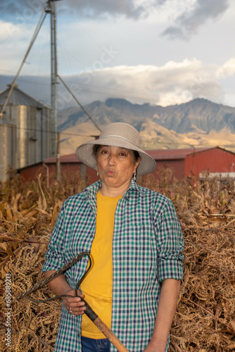 Mujer adulta retratada al altardecer en su granja lechera esta sosteniendo su rastrillo enfrente de un silo y una pila de hojas de maiz para el ganado  photo