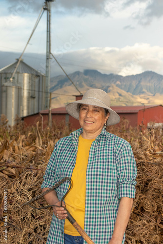 Retrato de un mujer adulta sonriendo al atardecer sosteniendo un rastrillo y esta vestida con sombrero de paja camisa a cuadros y de fondo sun silo un granero y las montañas de los andes en ecuador photo