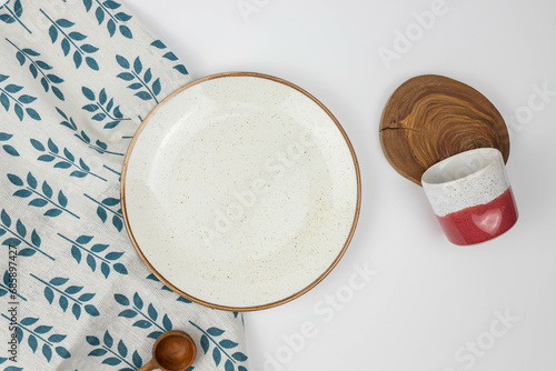 Composition of kitchen utilities  white plate  purple and white cup  wooden items on textile leaves pattern background  top view
