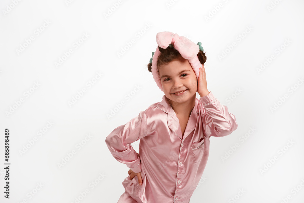 Cute little girl with curlers and a pink headband, wearing pink silk pajamas on a white background. Cute little girl with curlers and a pink headband, wearing pink silk pajamas on a white background.