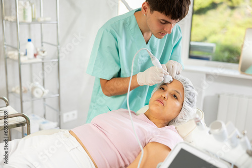 Young male cosmetologist performs facial ultrasound procedure with machine to young female client
