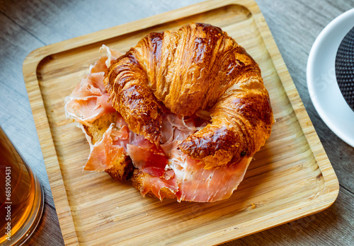Simple and satisfying snack is prepared for visitor of cafe - large croissant with slices of ham. Dish is served on wooden plate and optionally supplemented with glass of dark beer photo