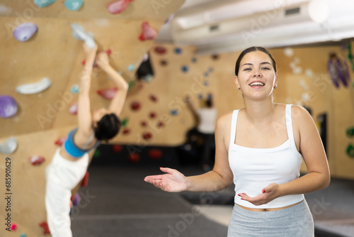 Positive slender europian girl in sportswear is standing near bouldering wall in gym. Clients of sports complex climb steep artificial wall in background photo