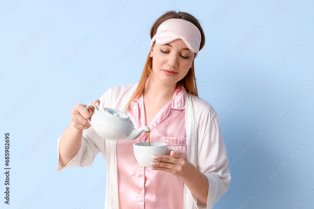 Young woman in pajamas pouring tea into cup on blue background Young woman in pajamas pouring tea into cup on blue background