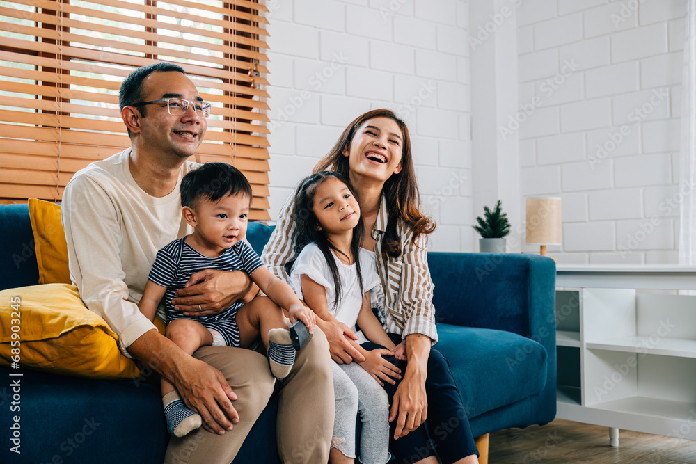 In the heart of their modern grooved house a joyful Asian family enjoys quality family time watching TV together on the cozy sofa. Laughter smiles and togetherness define their weekend. In the heart of their modern grooved house a joyful Asian family enjoys quality family time watching TV together on the cozy sofa. Laughter smiles and togetherness define their weekend.
