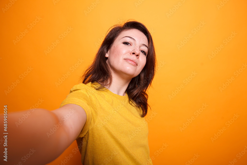 POV of caucasian female influencer wearing yellow t-shirt, taking selfie with camera while on isolated orange background. Brunette carefree vlogger smiling at camera while recording a daily vlog. POV of caucasian female influencer wearing yellow t-shirt, taking selfie with camera while on isolated orange background. Brunette carefree vlogger smiling at camera while recording a daily vlog.