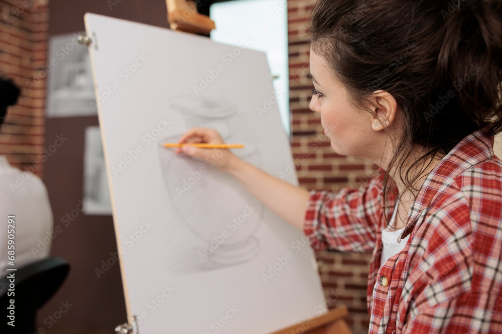 Young woman sitting at easel sketching basic vase on canvas using pencil, student girl learning still life basics while studying in art school, people drawing to relax and relieve stress Young woman sitting at easel sketching basic vase on canvas using pencil, student girl learning still life basics while studying in art school, people drawing to relax and relieve stress
