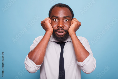 Quadro su tela Over a blue background, a nervous entrepreneur with open eyes holds his face in his closed palms