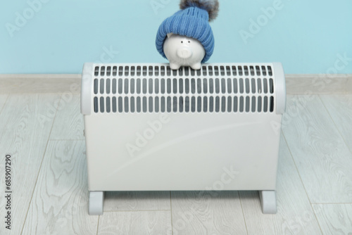 Electric convector heater, piggy bank and hat near blue wall. Heating season photo