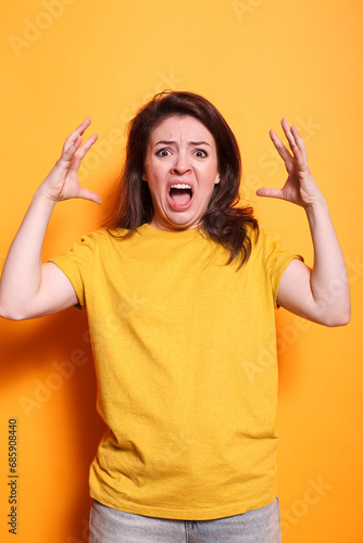 Caucasian woman with open mouth and raised arms, conveying anger and negativity. Annoyed lady in casual clothing stands in studio with orange background, screaming at the camera.