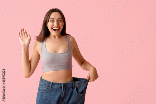 Beautiful young woman in loose jeans waving hand on pink background. Weight loss concept © Pixel-Shot