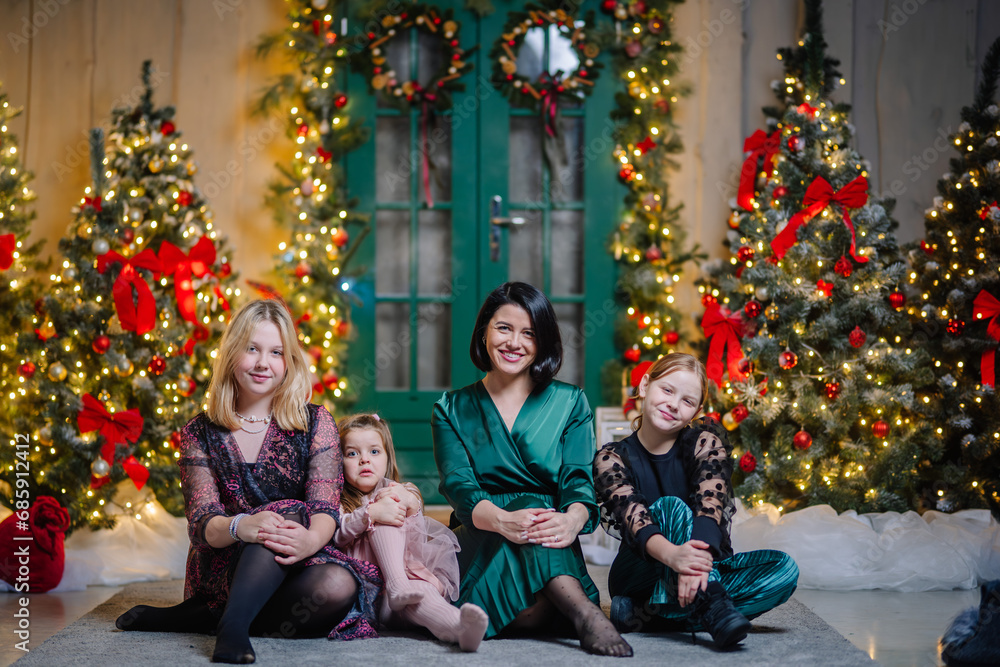 Mother with three daughters sitting and laughing in Christmas decorations, near a green door and many Christmas trees