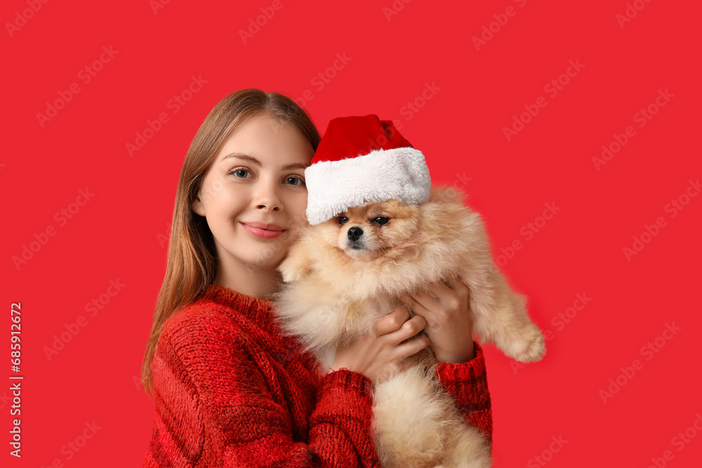 Young woman with cute Pomeranian dog in Santa hat on red background Young woman with cute Pomeranian dog in Santa hat on red background