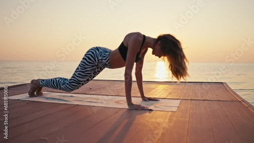 A young blonde girl in a black top in black and white pants does wave exercises while doing yoga and Cobra pose exercises on a wooden pier near the sea at Sunrise photo
