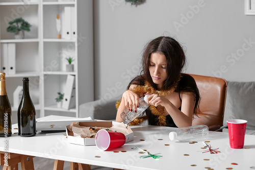 Young woman with hungover drinking water at table in office after New Year party photo