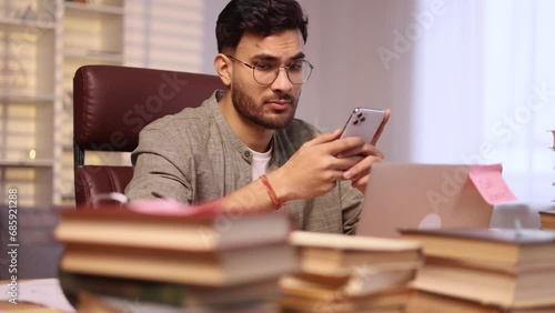 Boring exhausted young man student or freelancer scrolling feed of social networks products in internet store on smartphone instead of work or education at home workplace Procrastination concept photo