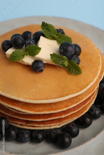 Stack of tasty pancakes with blueberries, butter and mint on light blue background, closeup