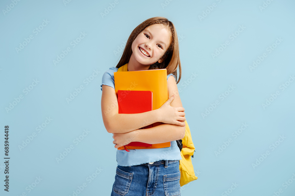 Portrait smiling cute school girl holding book, notebook, wearing backpack looking at camera isolated on blue background. Education, back to school concept Portrait smiling cute school girl holding book, notebook, wearing backpack looking at camera isolated on blue background. Education, back to school concept