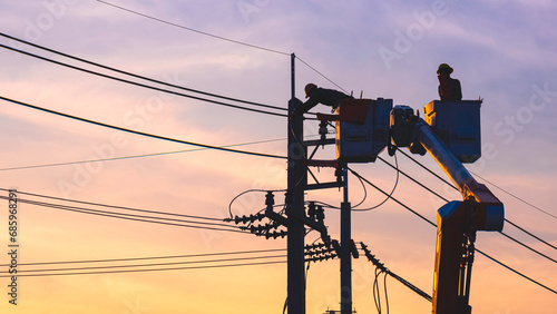 Silhouette of 2 electricians on bucket boom truck are repairing electrical system on electric power pole against sunset sky background in evening time
