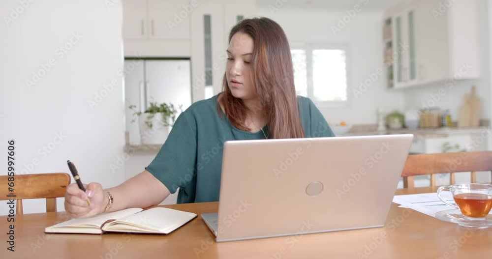 Busy plus size biracial woman writing notes and using laptop, working from home, slow motion Busy plus size biracial woman writing notes and using laptop, working from home, slow motion
