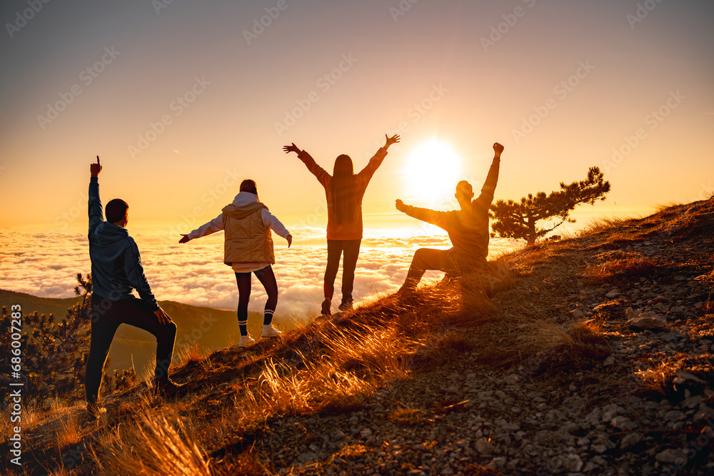 Group of happy friends are standing at mountain top and greeting sunrise or sunset above clouds - obrazy, fototapety, plakaty 