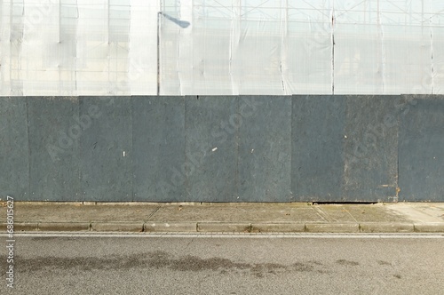 Black wooden panels fence with plastic sheet on scafolding covering building under construction . Concrete sidewalk and asphalt road in front. Background for copy space photo